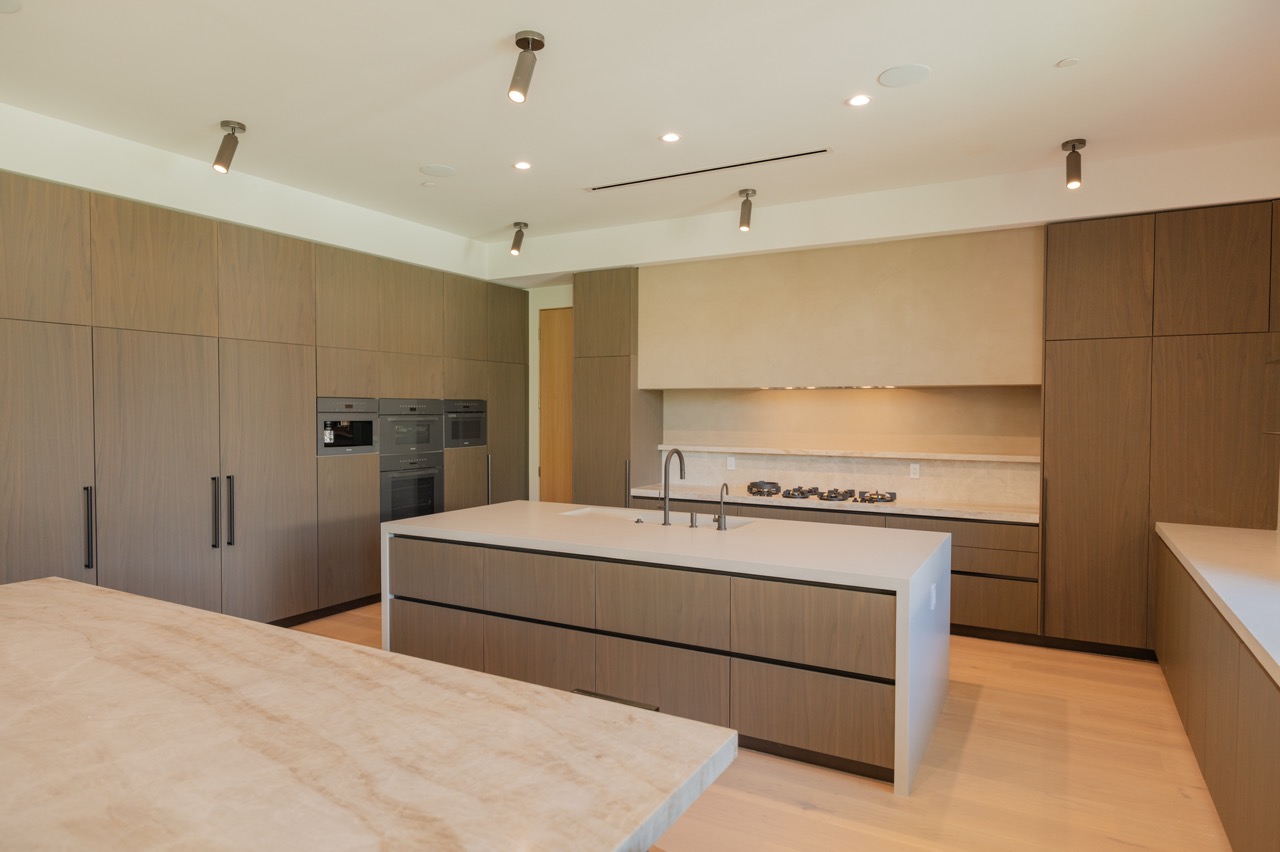 Wide walnut frameless kitchen with a plaster hood, travertine backsplash, integrated appliance wall, and a stone-topped island